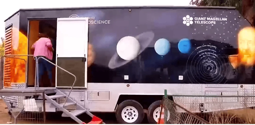 Tráiler móvil del Giant Magellan Telescope decorado con ilustraciones de planetas del sistema solar, con una persona subiendo las escaleras de acceso.