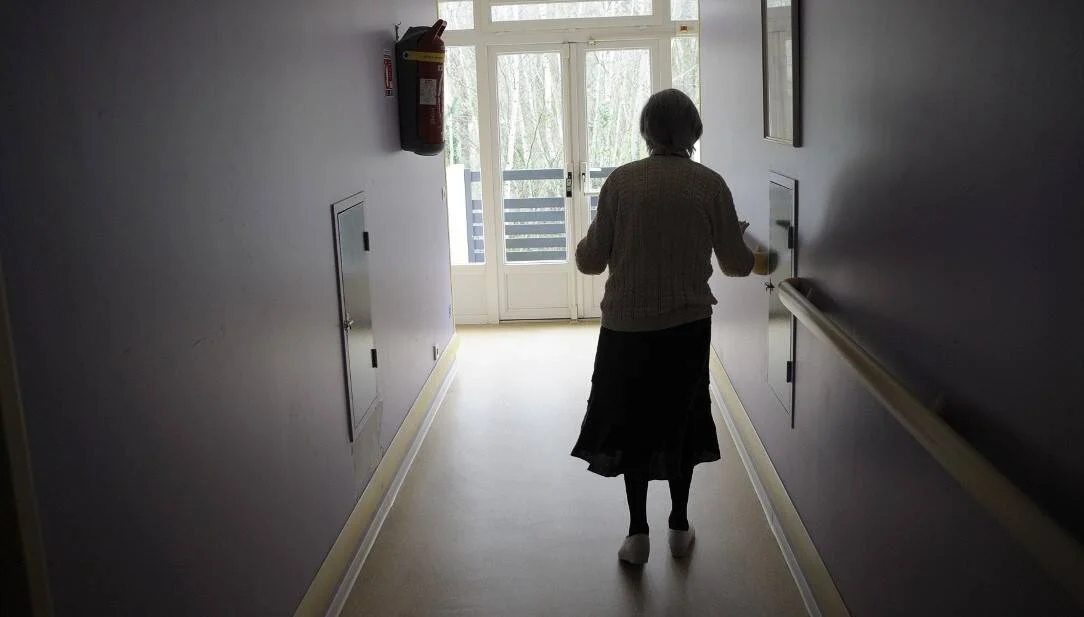 Anciana camina sola por el pasillo de una residencia geriátrica, apoyándose en la barandilla hacia una puerta con luz natural.