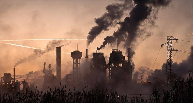 Complejo industrial con múltiples chimeneas expulsando humo negro y vapor en un cielo de tonos naranjas al atardecer.