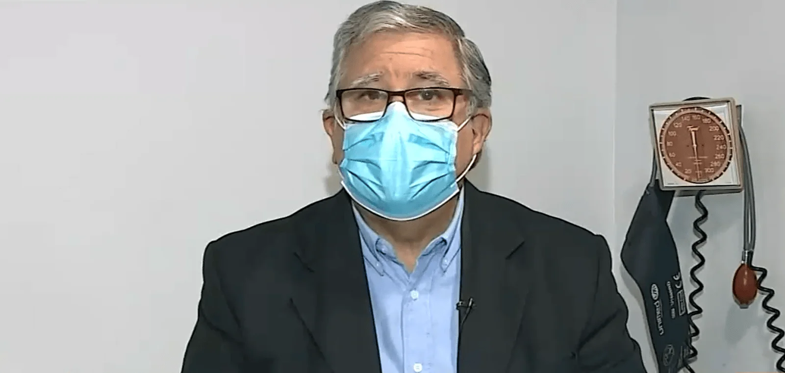 Hombre mayor con anteojos y mascarilla quirúrgica azul frente a un tensiómetro de pared en consultorio médico.