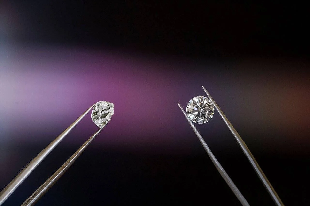 Dos diamantes de cortes distintos sostenidos individualmente con pinzas metálicas sobre fondo oscuro con luz violeta.