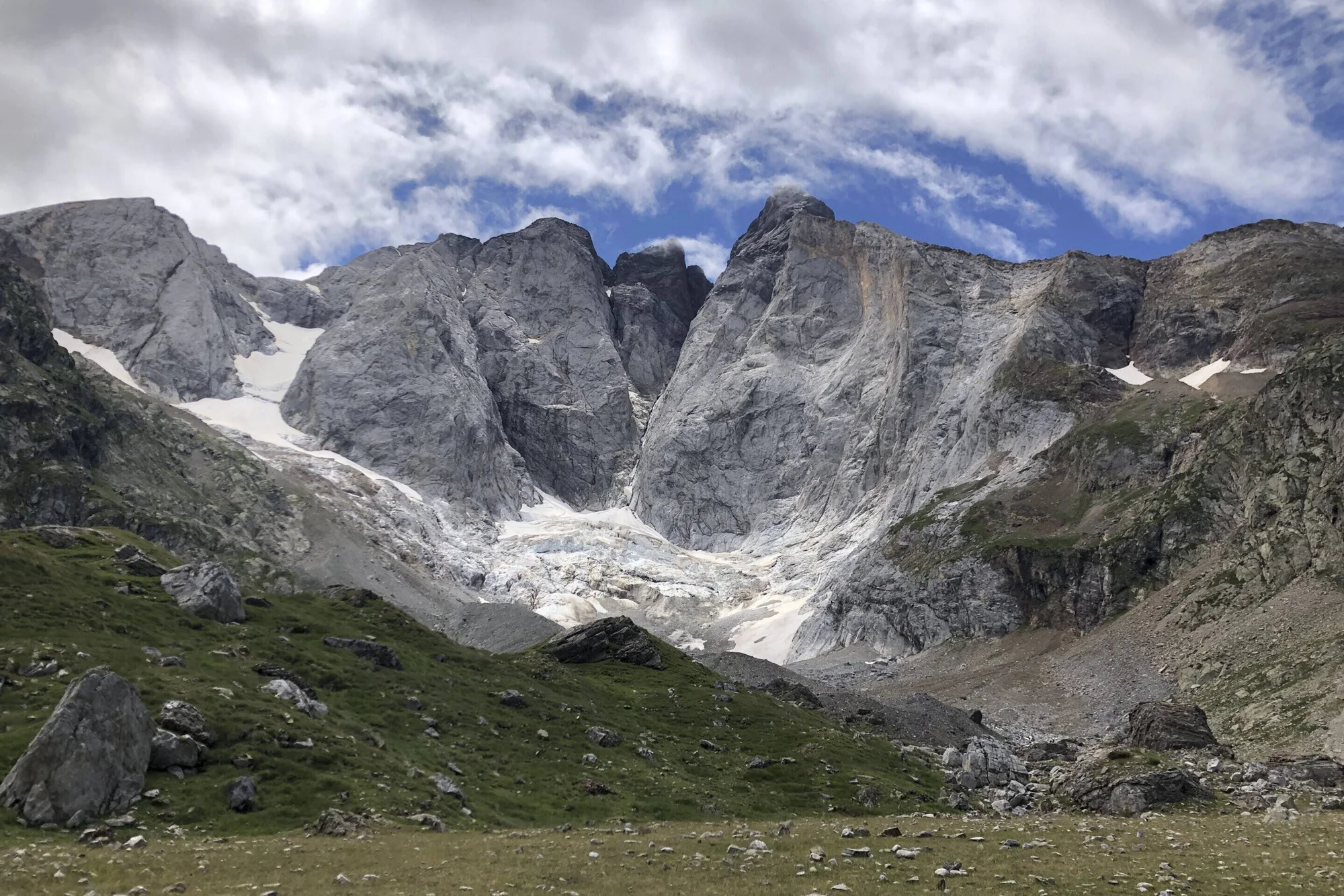 Circo glaciar de alta montaña con paredes rocosas escarpadas, restos de nieve y hielo en la base, y praderas verdes en primer plano.