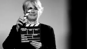 Mujer con cabello corto rubio sostiene una claqueta de filmación frente a su rostro, en foto en blanco y negro.