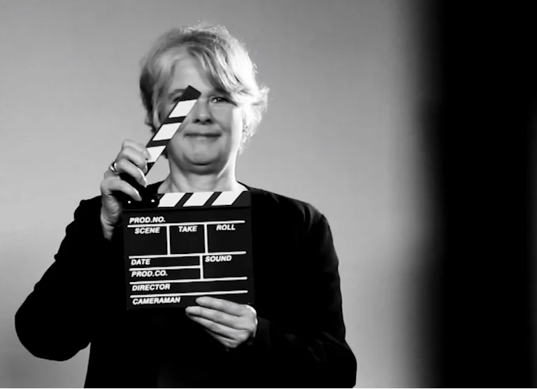 Mujer con cabello corto rubio sostiene una claqueta de filmación frente a su rostro, en foto en blanco y negro.