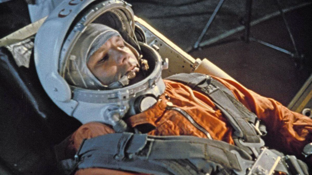 Cosmonauta con traje espacial naranja y casco abierto recostado en el asiento de una cápsula espacial soviética.