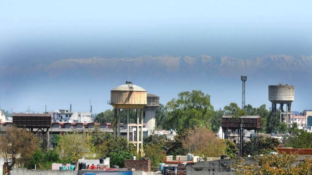 Panorama urbano con torres de agua elevadas, edificios y árboles, con una cadena montañosa envuelta en neblina al fondo.