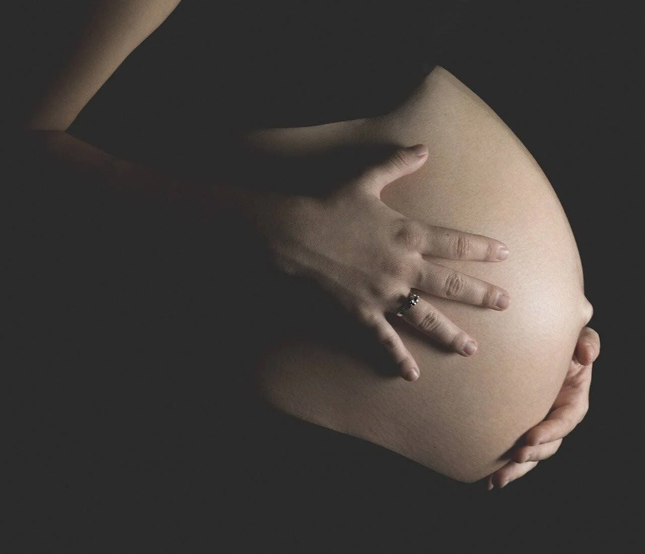 Mujer embarazada sostiene su vientre con ambas manos sobre fondo oscuro.