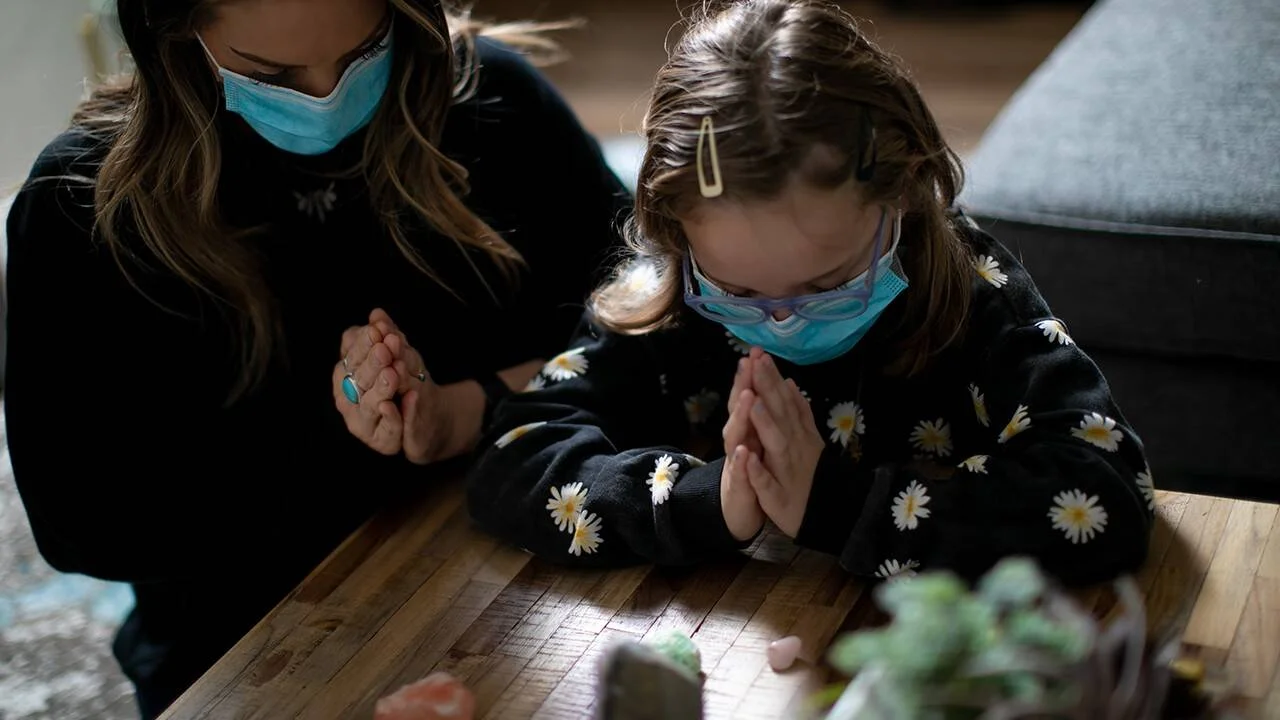 Mujer adulta y niña con mascarillas quirúrgicas rezan con manos juntas sobre una mesa de madera en un interior.
