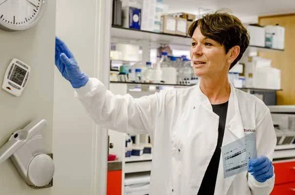 Mujer con bata blanca y guantes de látex sostiene una muestra en un laboratorio de toxicología.