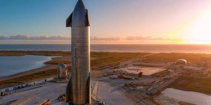 Prototipo de cohete Starship de acero inoxidable erguido en la base de lanzamiento junto a la costa al atardecer.