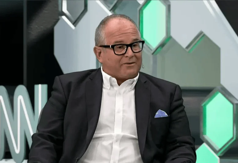 Hombre de cabello canoso con anteojos negros y traje oscuro sentado en un estudio de televisión con fondo geométrico verde y gris.