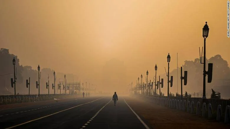 Una persona camina sola por una amplia avenida envuelta en densa neblina de contaminación bajo un cielo anaranjado.