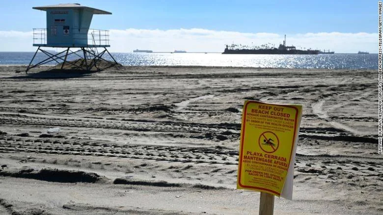 Cartel amarillo de playa cerrada por agua contaminada frente a una torre de vigilancia y barcos en el horizonte.