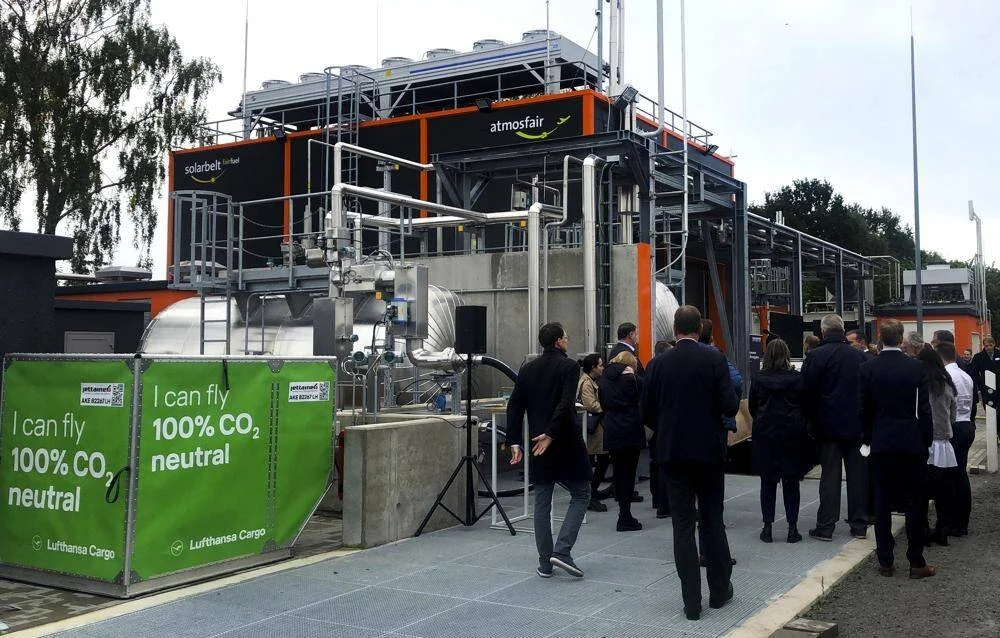 Grupo de personas visita una planta industrial de Atmosfair con carteles de Lufthansa Cargo sobre vuelos 100% neutros en CO₂.