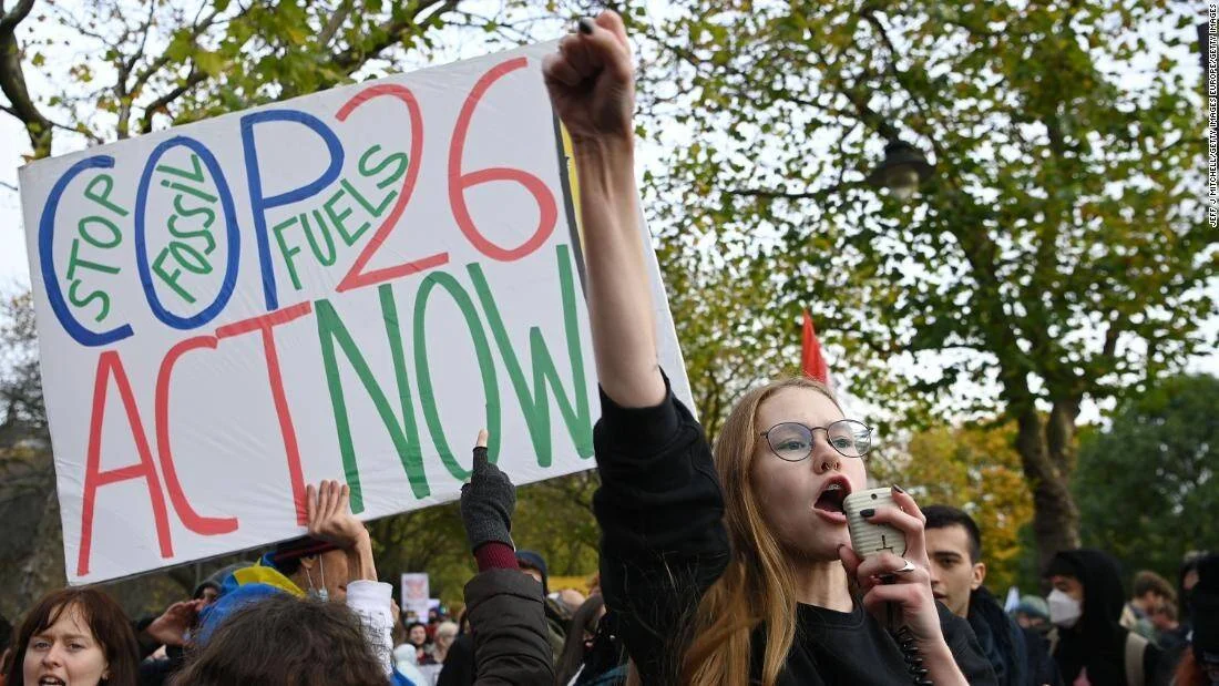Joven activista climática con megáfono y puño en alto junto a cartel que dice 'Stop Fossil Fuels COP26 Act Now' en una manifestación.