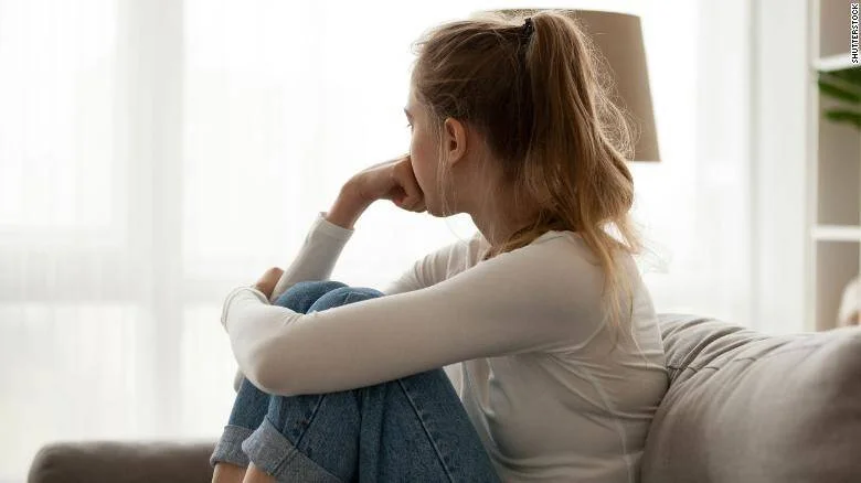 Joven sentada en un sofá con las rodillas al pecho, mirando hacia la ventana en actitud pensativa o triste.