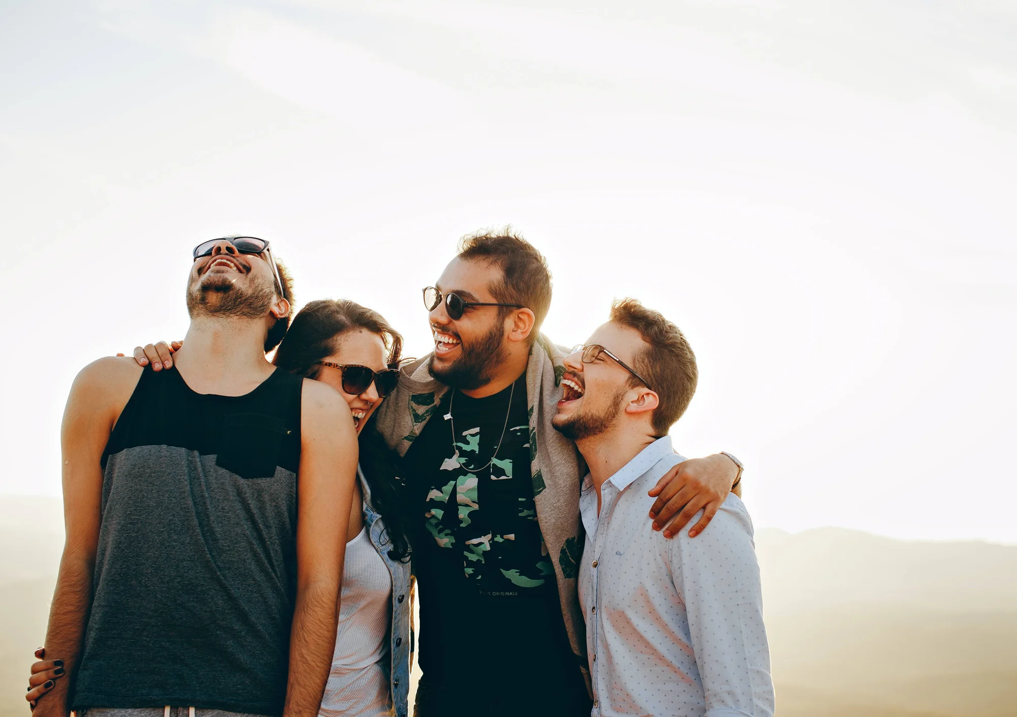 Cuatro jóvenes amigos abrazados ríen juntos al aire libre bajo un cielo despejado.