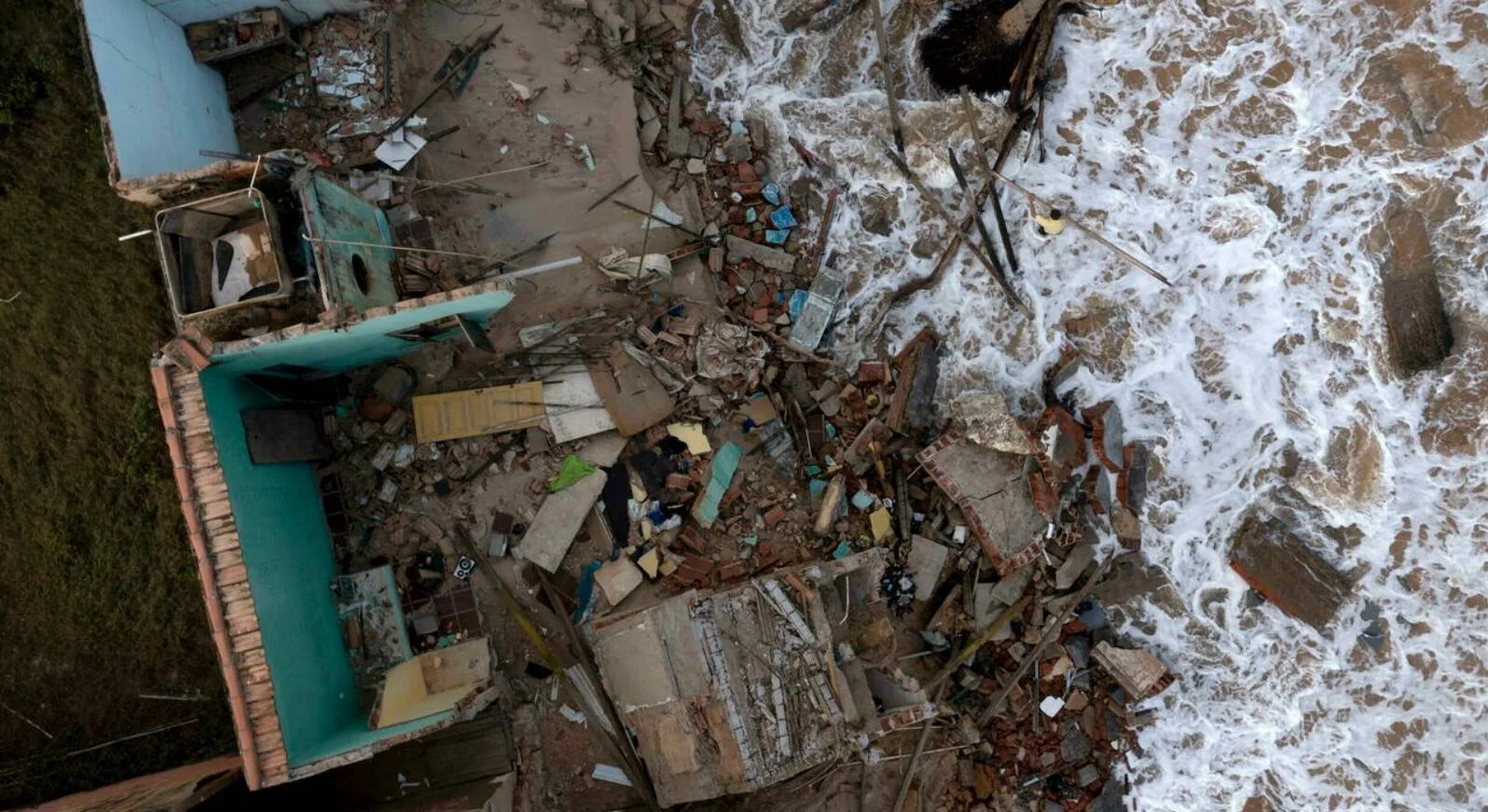 Fotografía aérea de una casa completamente demolida en la orilla del mar, con escombros y olas rompiendo sobre los restos.
