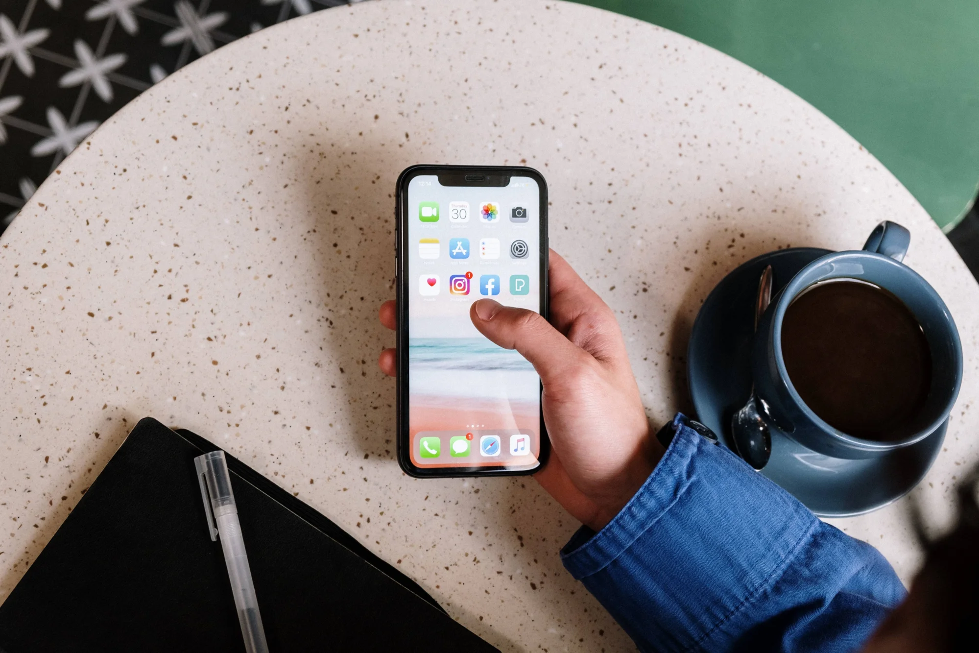 Una mano sostiene un iPhone mostrando la pantalla de inicio con íconos de Instagram y Facebook, junto a una taza de café sobre una mesa.