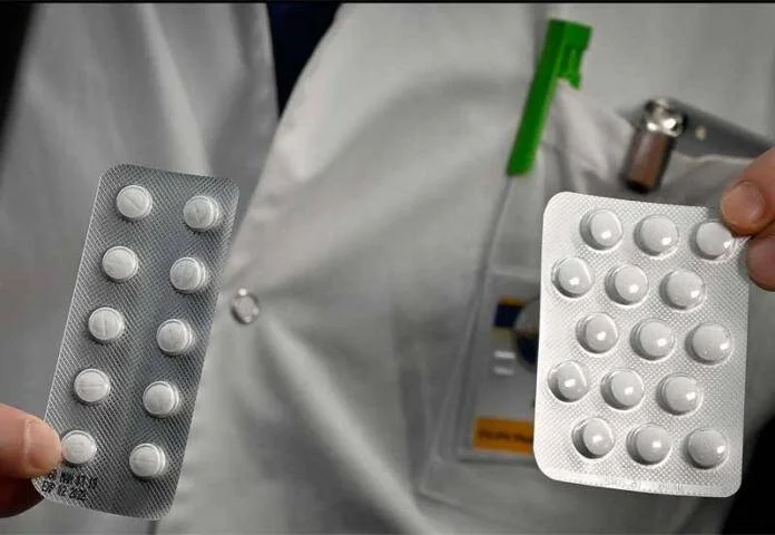 Persona con guardapolvo blanco sostiene dos blisteres de comprimidos blancos, uno en cada mano.