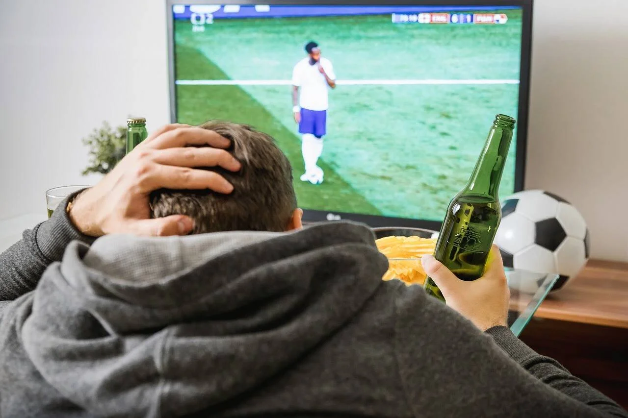 Hombre con la mano en la cabeza mira un partido de fútbol por TV, sosteniendo una cerveza y con papas fritas a su lado.