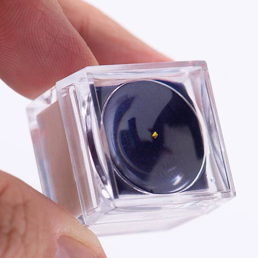 Mano sosteniendo una caja de plástico transparente con lupa que muestra una minúscula pepita de oro sobre fondo negro.