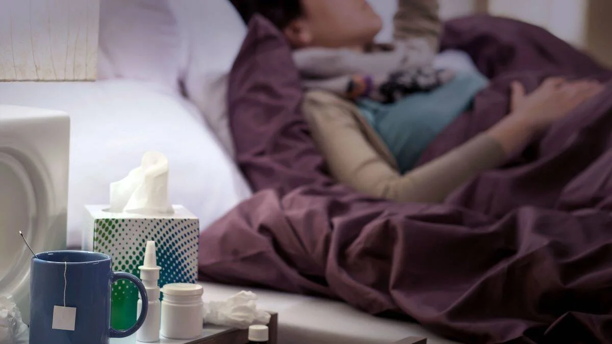 Persona acostada en cama con fiebre, junto a una mesita con té, pañuelos y medicamentos para aliviar síntomas de gripe.