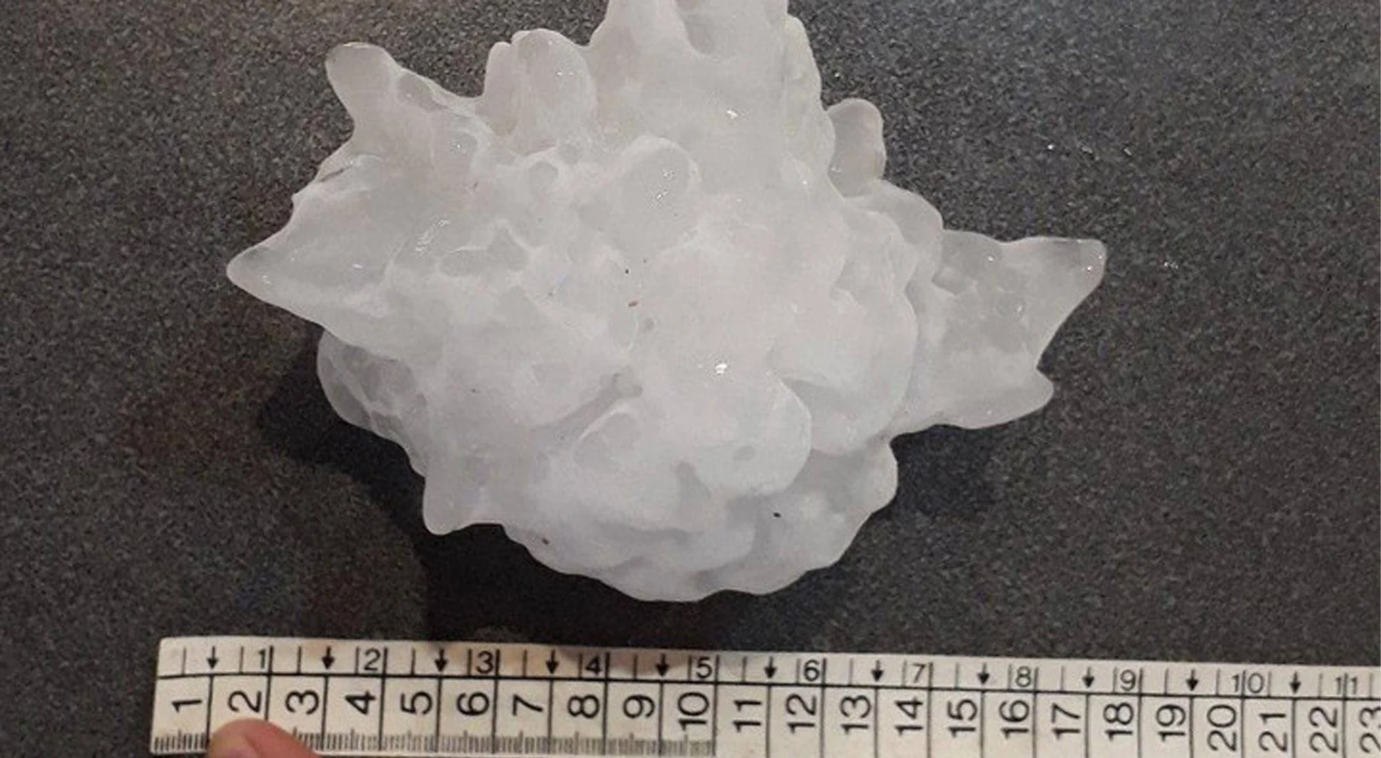 Piedra de granizo de gran tamaño sobre superficie gris, medida con una regla que indica aproximadamente 10 cm de diámetro.