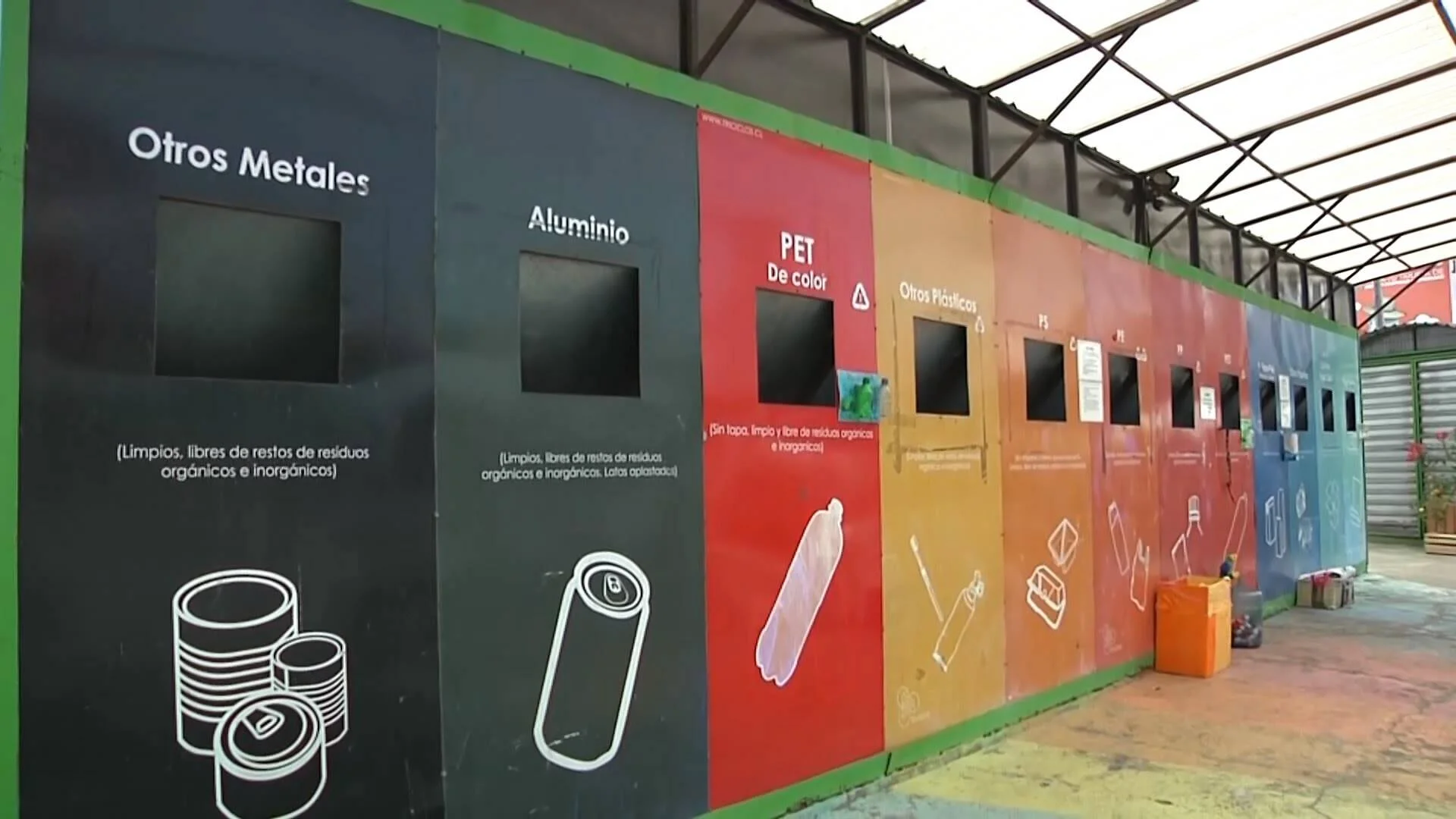 Contenedores de reciclaje clasificados por material: otros metales, aluminio, PET de color, otros plásticos y PS, en un punto limpio techado.