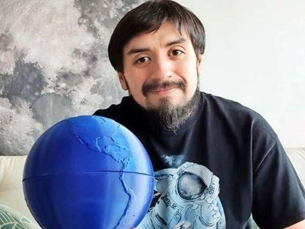 Hombre joven con barba sostiene un globo terráqueo azul hecho a mano, con un mural de la Luna de fondo.