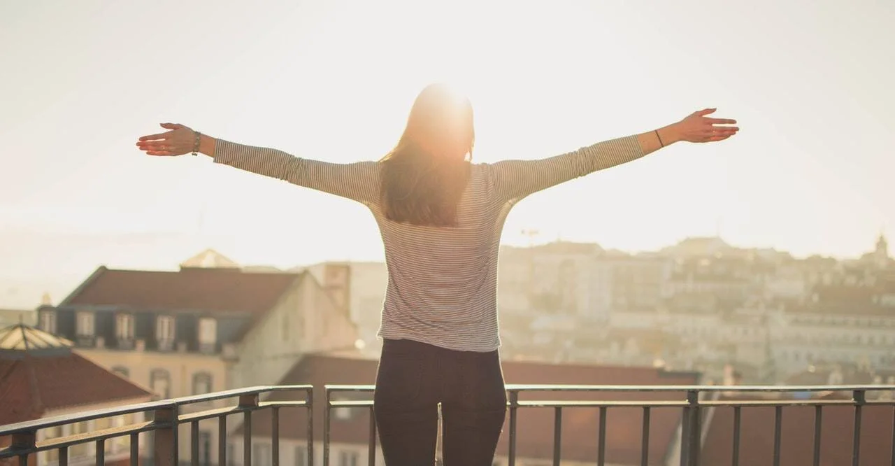 Mujer de espaldas con los brazos extendidos en una terraza, contemplando un paisaje urbano bañado por el sol.