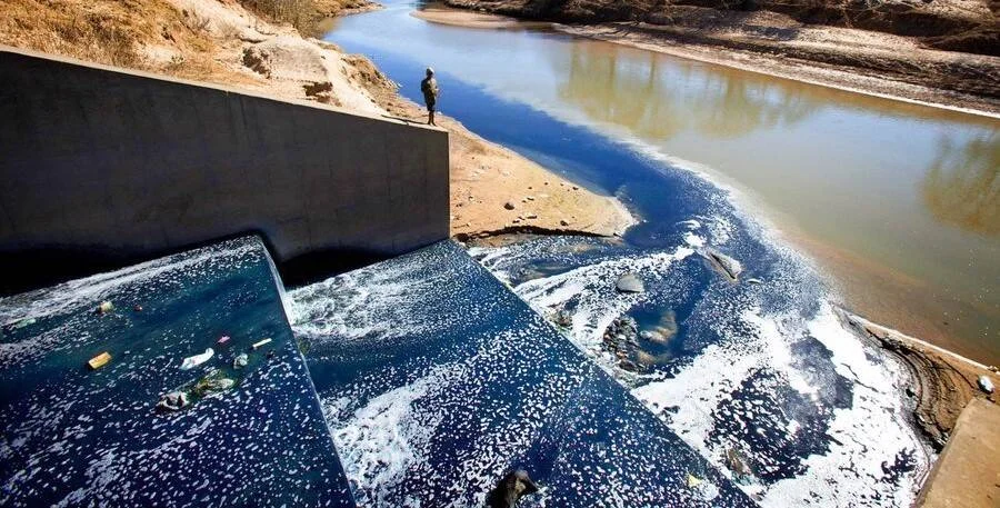 Agua contaminada y espumosa de color oscuro descarga desde una estructura de concreto hacia un río, mientras una persona observa desde la orilla.