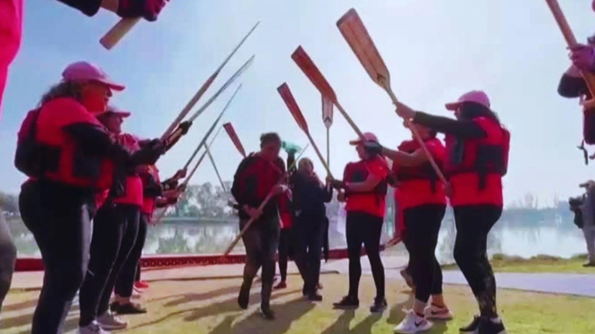 Grupo de mujeres con uniformes rojos y salvavidas forman túnel de remos al aire libre junto a un lago.