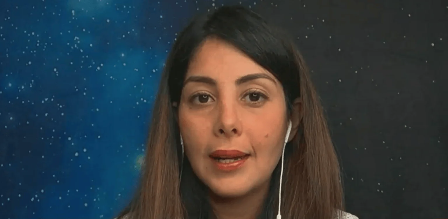 Mujer de cabello castaño con auriculares blancos habla frente a un fondo con imagen del espacio estrellado.