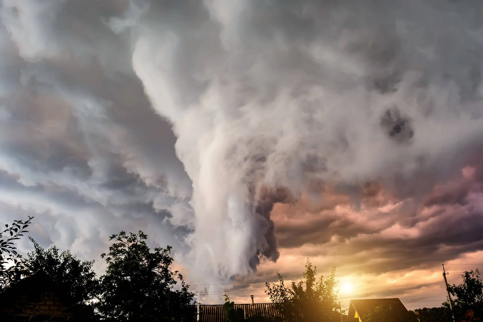 Un tornado de gran magnitud desciende entre nubes tormentosas sobre tejados y árboles al atardecer.
