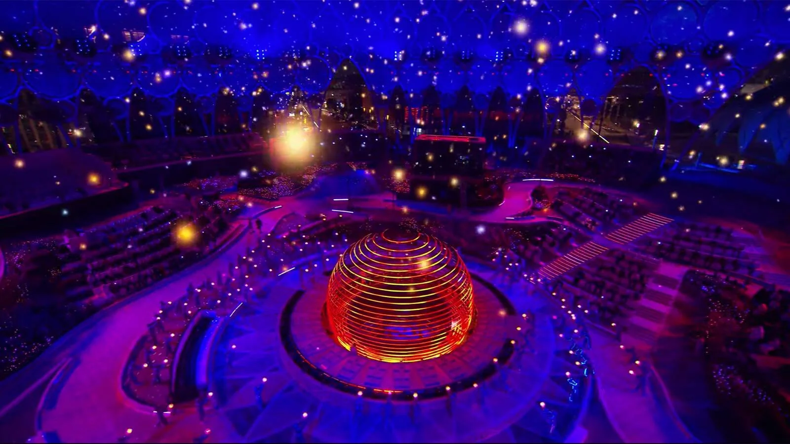 Vista aérea de una ceremonia con una gran esfera naranja iluminada en el centro de un estadio bajo un cielo azul estrellado.