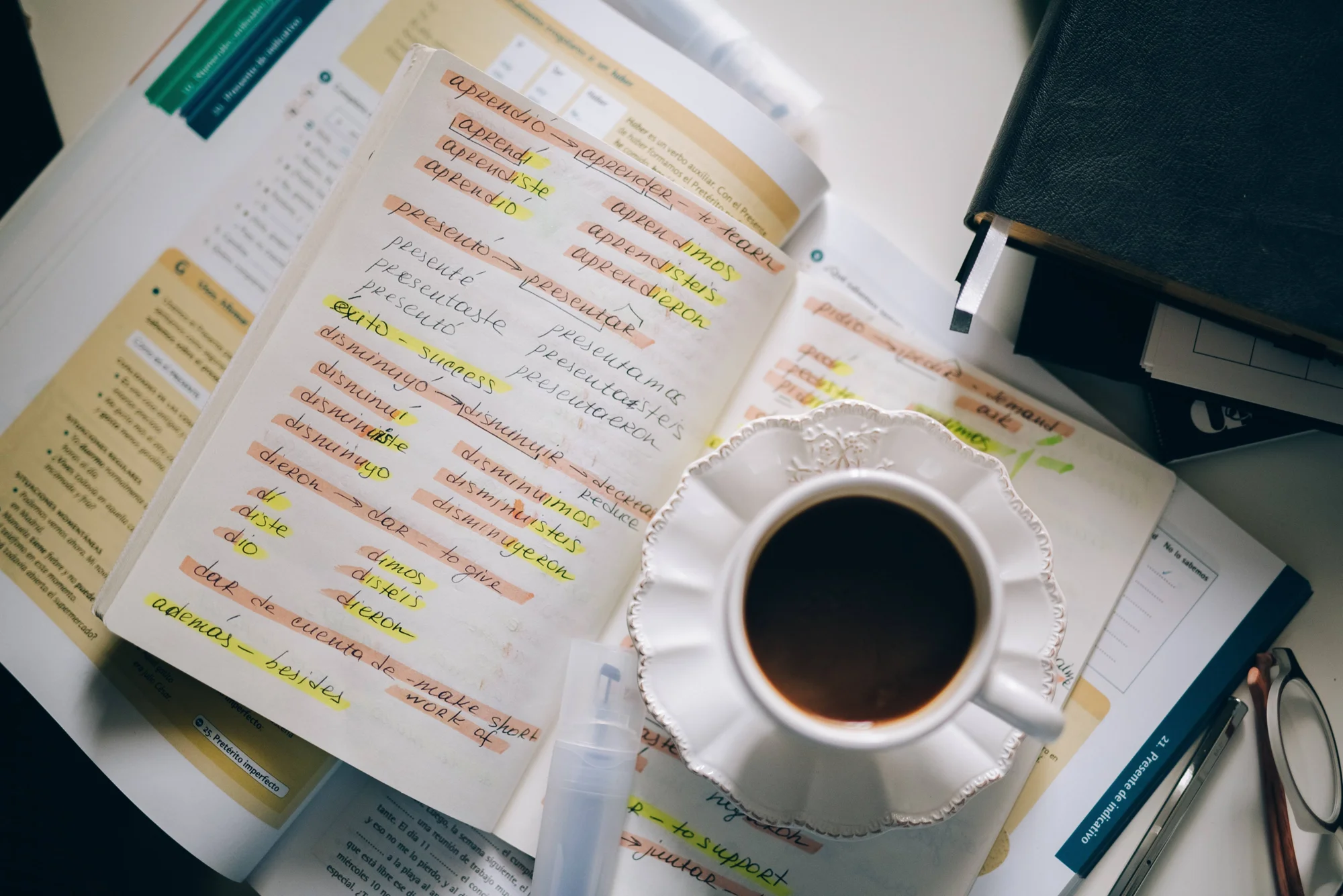 Cuaderno con conjugaciones de verbos en español resaltadas, junto a una taza de café y libros de gramática sobre una mesa de estudio.