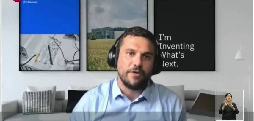 Hombre con auriculares habla en videollamada desde su hogar, con cuadros decorativos y texto 'I'm Inventing What's Next' al fondo.