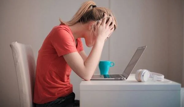 Mujer con remera roja sostiene su cabeza con ambas manos frente a una laptop, en actitud de estrés o agotamiento.