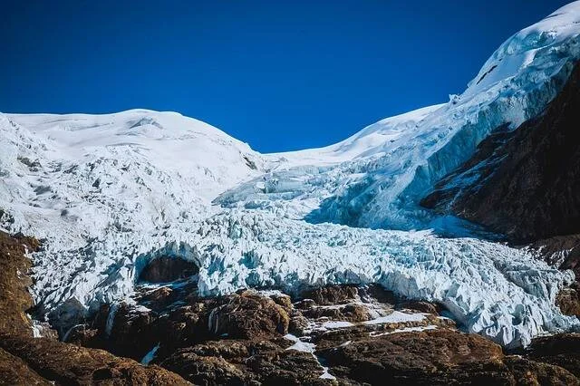 Glaciar con seracs de hielo azulado desciende entre montañas nevadas bajo un cielo azul despejado.