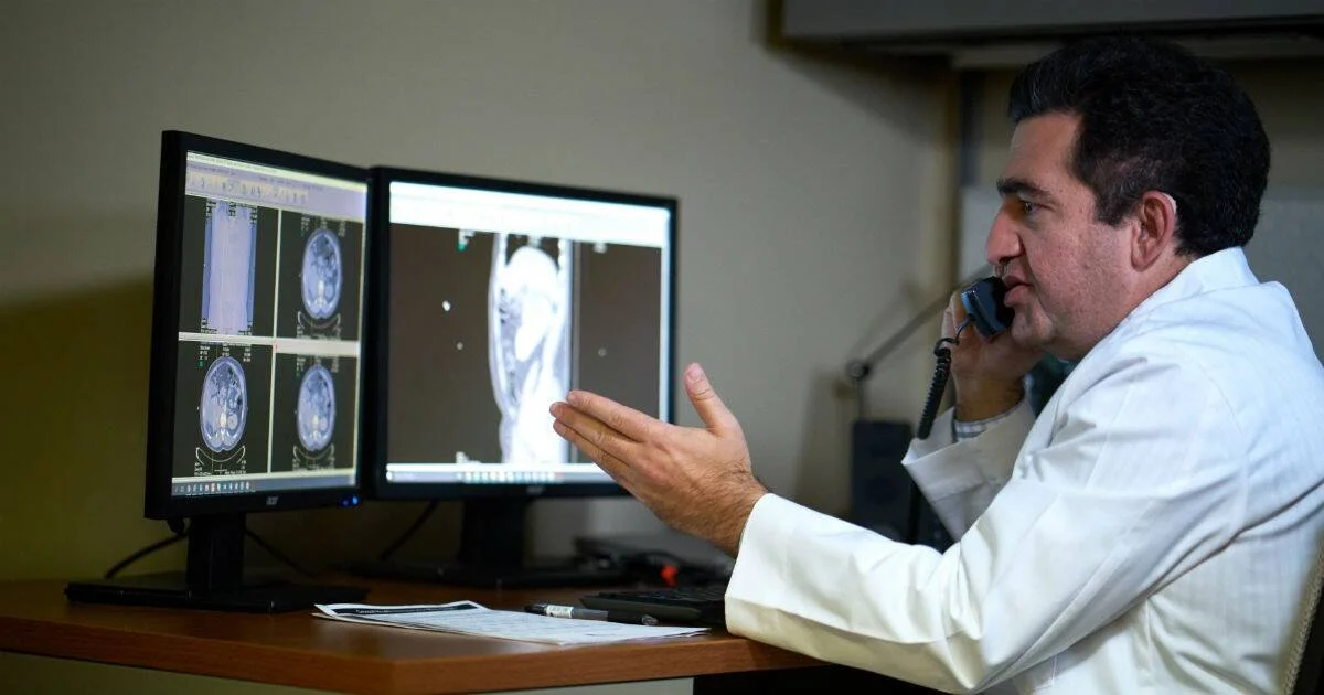 Médico con guardapolvo blanco habla por teléfono frente a dos monitores que muestran imágenes de tomografía computada.