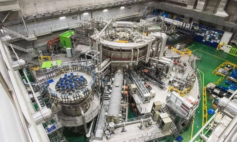 Vista aérea del interior de una instalación de fusión nuclear con un gran reactor tokamak rodeado de tuberías, cables y estructuras metálicas industriales.