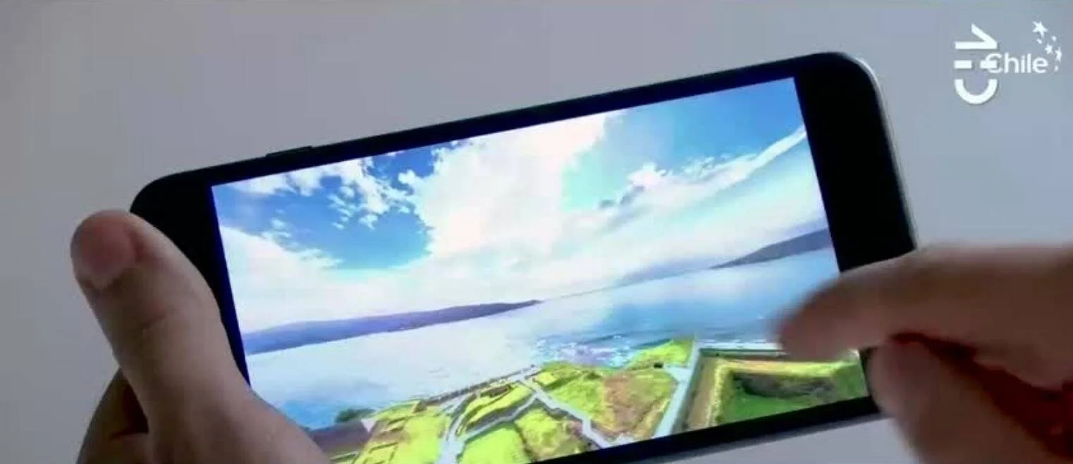 Persona sostiene un smartphone en horizontal mostrando una vista aérea de una zona costera verde con cielo azul.