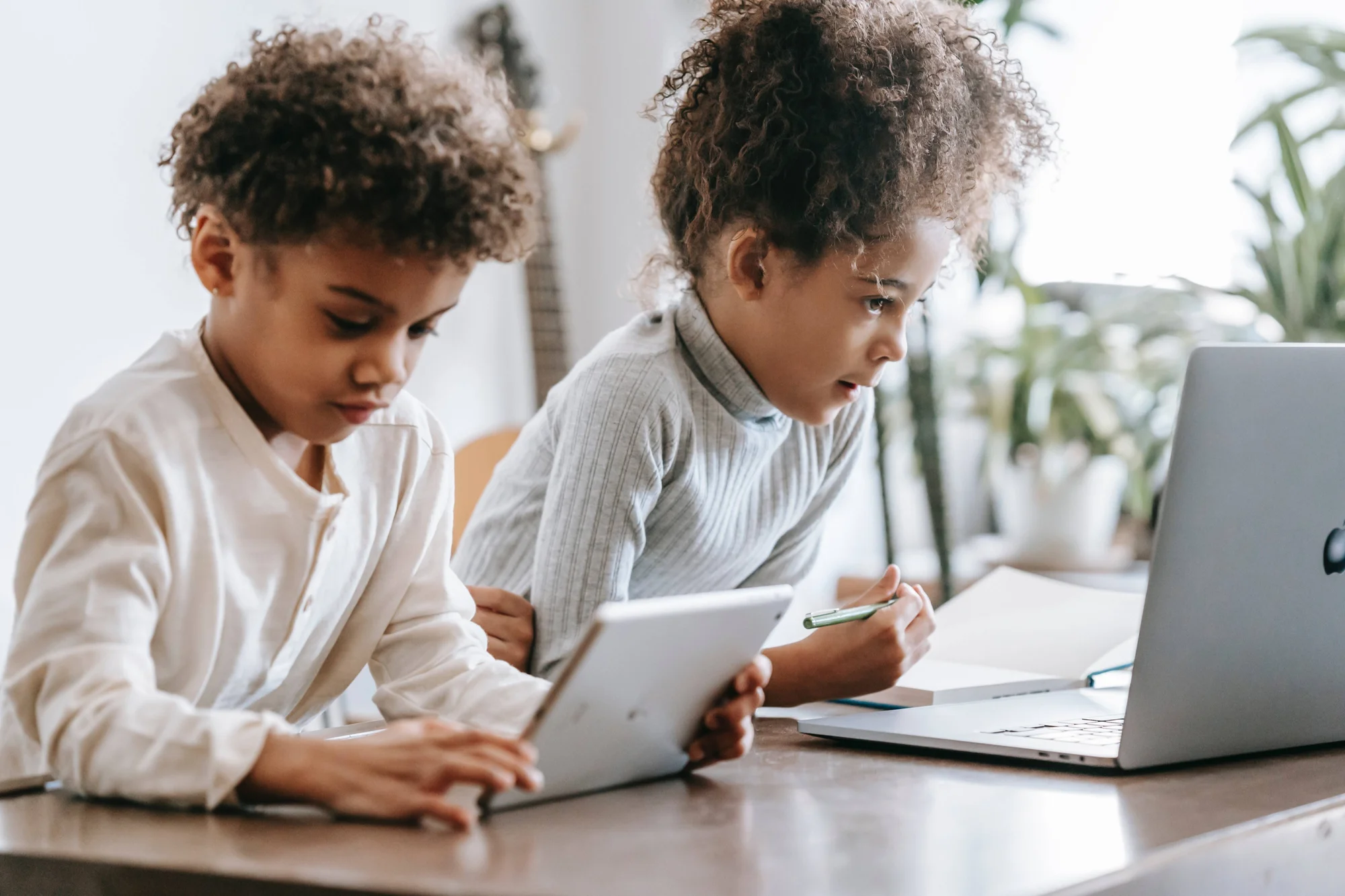 Dos niños con cabello rizado usan una tablet y una laptop juntos mientras estudian en una mesa.