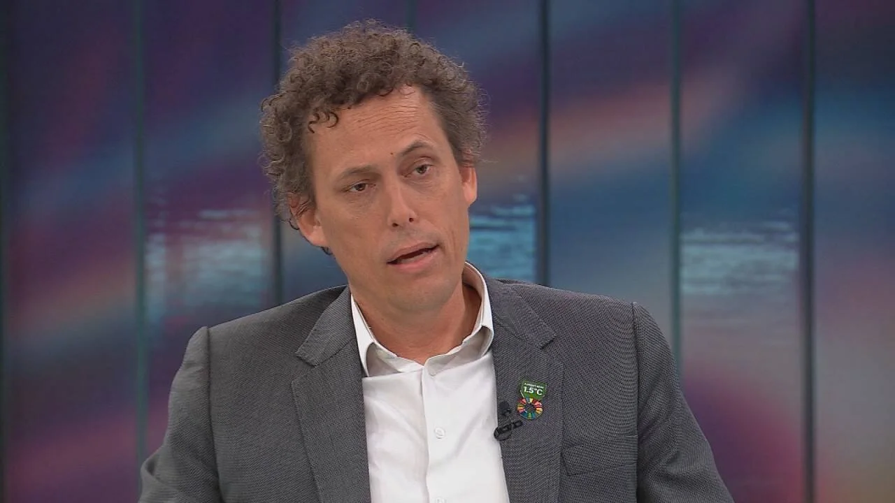 Hombre de cabello rizado con blazer gris y camisa blanca habla en estudio de televisión; lleva un pin de los Objetivos de Desarrollo Sostenible con la leyenda 1,5 °C.