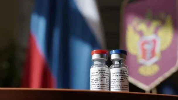 Dos viales de vacuna con tapas roja y azul frente a la bandera de Rusia y el escudo del Ministerio de Salud.