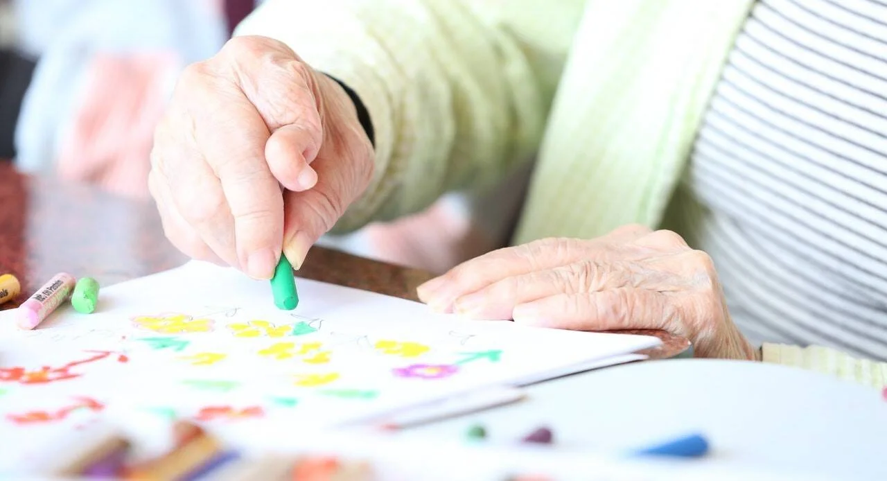 Manos ancianas colorean flores con crayones sobre papel en una actividad recreativa.