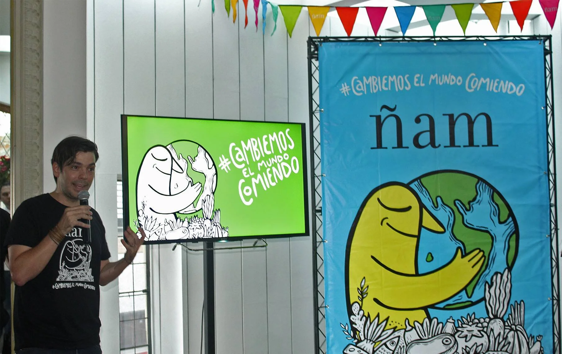 Un hombre con micrófono presenta el festival gastronómico Ñam junto a una pantalla y un banner con el lema #CambiemosElMundoComiendo.