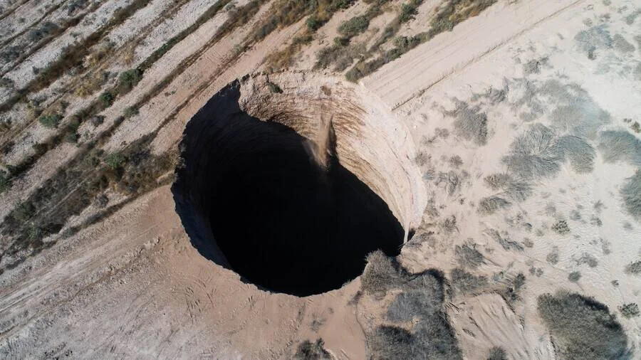 Vista aérea de un enorme socavón de paredes oscuras abierto en un suelo árido y seco rodeado de vegetación escasa.
