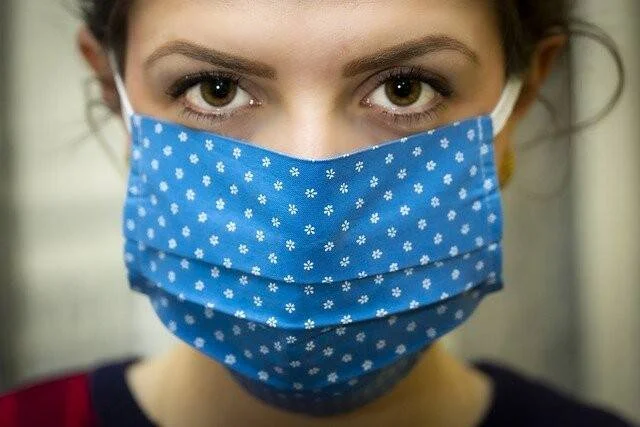 Mujer joven con mascarilla de tela azul con flores blancas mirando a cámara.